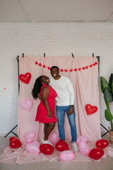 Young African couple embracing while standing on pink fabric background with heart shaped balloons