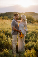 Happy and in love pregnant couple with a bouquet of sunflowers spending time together outdoors at sunset.