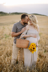 Portrait of a happy pregnant couple with a bouquet of sunflowers spending time together walking in a wheat field at sunset