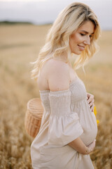 Fototapeta na wymiar Portrait of a happy pregnant blonde woman with a straw basket and a bouquet of sunflowers spending time outdoors in a wheat field at sunset. The concept of motherhood.