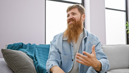 Fototapeta premium Cheerful young redhead man confidently speaking, relaxing on a cozy sofa at home, exuding happiness while sitting, a radiant smile gracing his handsome face