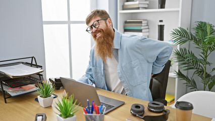 Young redhead guy with glasses suffering backache, anxiously touching injured spine while working indoors, sitting at the office table