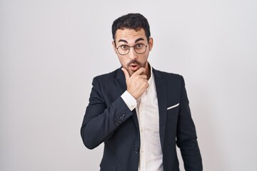 Handsome business hispanic man standing over white background looking fascinated with disbelief, surprise and amazed expression with hands on chin
