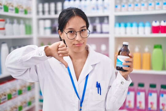 Young hispanic woman working at pharmacy drugstore holding syrup with angry face, negative sign showing dislike with thumbs down, rejection concept