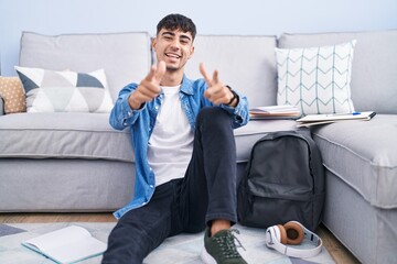 Young hispanic man sitting on the floor studying for university pointing fingers to camera with...