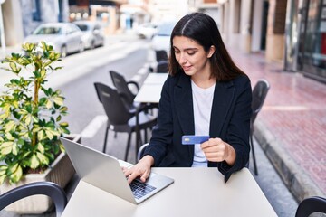 Young beautiful hispanic woman using laptop and credit card sitting on table at coffee shop terrace