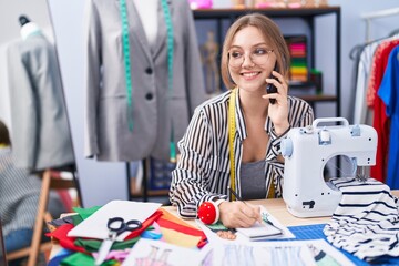 Young blonde woman tailor talking on smartphone writing on notebook at tailor shop