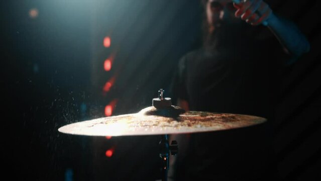 The drummer plays cymbal in a dark studio with lights at the back.