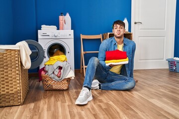 Young hispanic man holding clean laundry puffing cheeks with funny face. mouth inflated with air, catching air. - obrazy, fototapety, plakaty