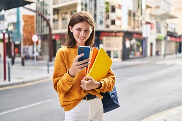 Young blonde woman student using smartphone holding books at street