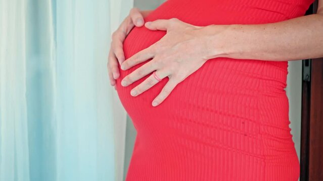 A young pregnant girl with a large stomach, stands at home in a colorful dress and touches her stomach with her hands.