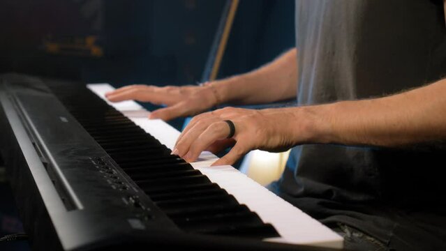 The young musician's hands play the piano in a dark studio with a light at the back.