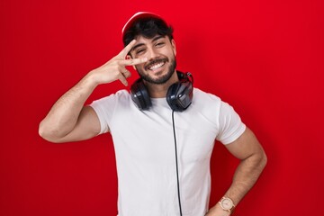 Hispanic man with beard wearing gamer hat and headphones doing peace symbol with fingers over face, smiling cheerful showing victory