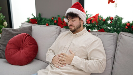 Young hispanic man suffering stomach ache wearing christmas hat at home