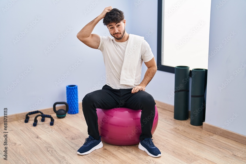 Canvas Prints Hispanic man with beard sitting on pilate balls at yoga room confuse and wonder about question. uncertain with doubt, thinking with hand on head. pensive concept.