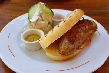 Dish of a sandwich of a pork and beef sausage in toasted bread, potato and cucumber salad and a mustard sauce on the side