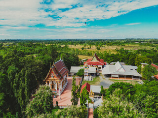 High angle photo, taken with a drone, landscape of the Thai temple area, surrounded by trees, bright sky, lots of sunshine.