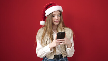 Young blonde woman wearing christmas hat using smartphone over isolated red background