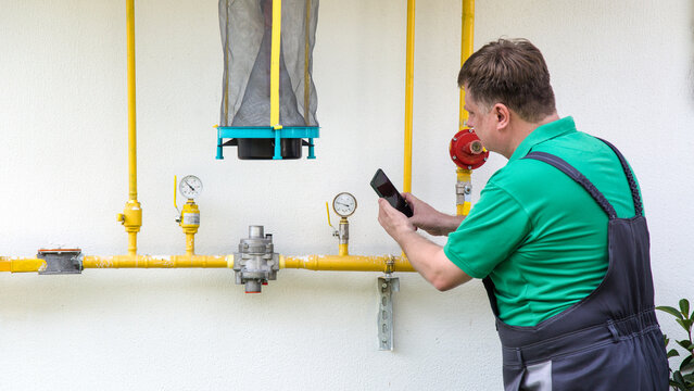 A man in work clothes looks and takes photographs water and gas consumption meters on a smartphone.