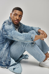 good looking young african american man in trendy denim outfit sitting on floor, fashion concept