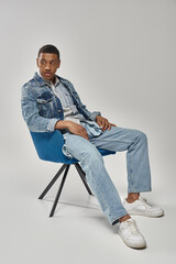 handsome african american man in stylish denim outfit sitting on blue chair, fashion concept