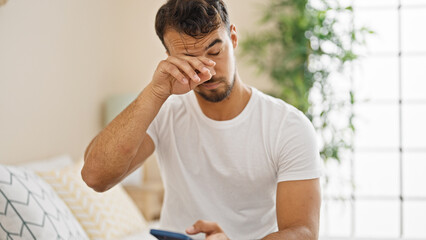 Young hispanic man waking up using smartphone at bedroom