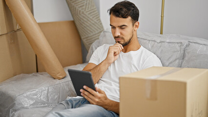 Young hispanic man using touchpad thinking at new home