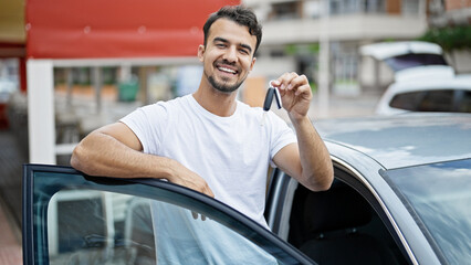 Young hispanic man holding key of new car leaning on door car at street