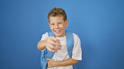 Adorable blond boy student, full of joy, laughing a lot while pointing to the camera against isolated blue wall background © Krakenimages.com