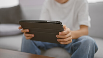 Adorable child comfortably sitting on sofa, hands using touchpad of a laptop relaxing at home in the living room, his eyes glued to the device, immersed in kid-friendly online content