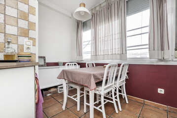 White wooden kitchen dining table with matching chairs, red tablecloth and brown stoneware floors under the window