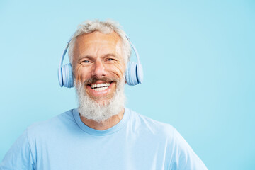 Happy handsome mature man, gray haired bearded hipster listening music in wireless headphones isolated on blue background. Positive lifestyle, technology concept