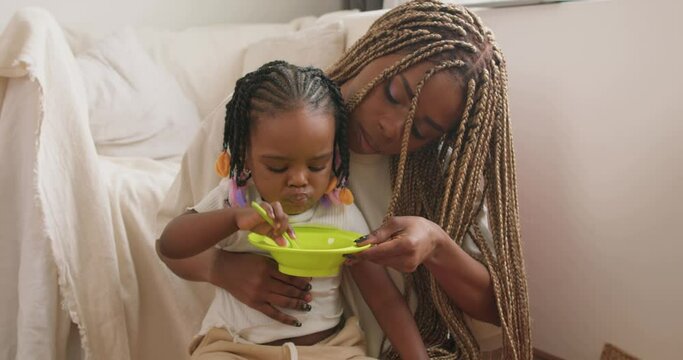 little nice adorable african kid learning to eat herself, mother holding a plate with porridge, slow motion,little daughter playing with food, dish health care, mother is against snacks and fast food