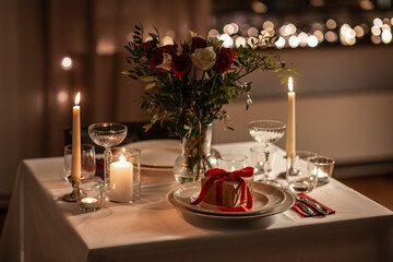 holidays, romantic date and celebration concept - close up of festive table serving for two with gift box, flowers in vase and candles burning at home on valentine's day at home