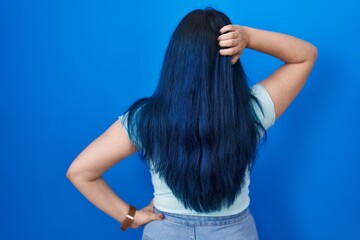 Young modern girl with blue hair standing over blue background backwards thinking about doubt with hand on head