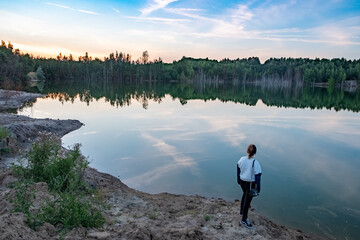 The image depicts a solitary figure standing at the edge of a tranquil lake, lost in contemplation as the day transitions to evening. The vastness of the water reflects the fading light and the