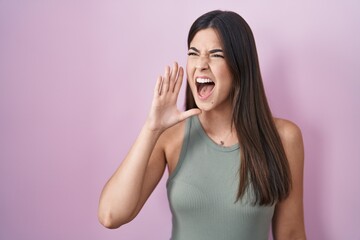 Hispanic woman standing over pink background shouting and screaming loud to side with hand on...