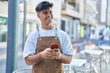 Young hispanic man waiter smiling confident using smartphone at coffee shop terrace