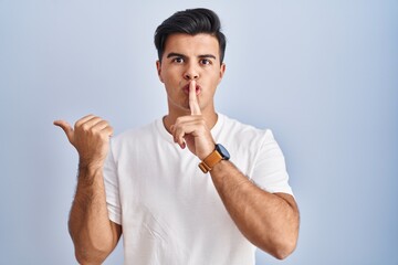 Hispanic man standing over blue background asking to be quiet with finger on lips pointing with...