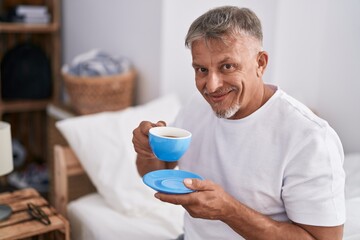 Middle age grey-haired man drinking cup of coffee sitting on bed at bedroom
