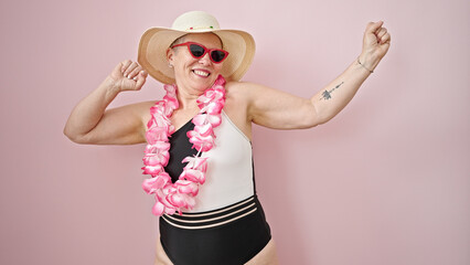 Middle age grey-haired woman tourist wearing swimsuit and hawaiian lei dancing over isolated pink...
