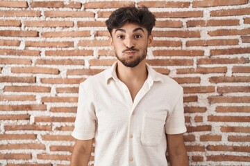 Arab man with beard standing over bricks wall background puffing cheeks with funny face. mouth...