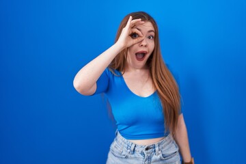 Redhead woman standing over blue background doing ok gesture shocked with surprised face, eye looking through fingers. unbelieving expression.