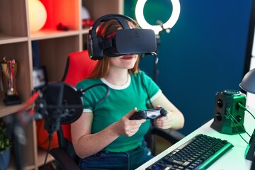 Young redhead woman streamer playing video game using virtual reality glasses and joystick at gaming room