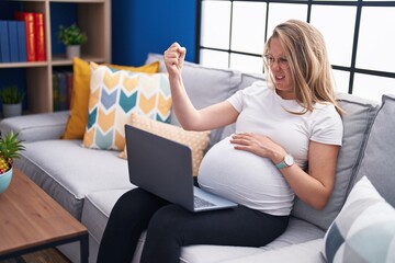 Young pregnant woman sitting on the sofa at home using laptop annoyed and frustrated shouting with anger, yelling crazy with anger and hand raised