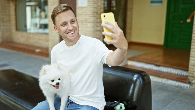 Cheerful, young caucasian man sitting on city street bench, joyfully taking a confident, happy selfie with his handsome pet dog using smartphone.