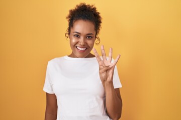 Young hispanic woman with curly hair standing over yellow background showing and pointing up with fingers number four while smiling confident and happy.
