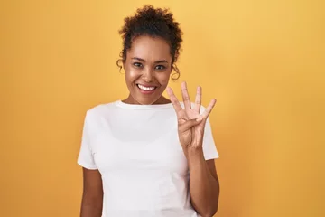 Fotobehang Young hispanic woman with curly hair standing over yellow background showing and pointing up with fingers number four while smiling confident and happy. © Krakenimages.com