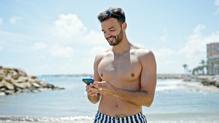 Young hispanic man tourist wearing swimsuit using smartphone smiling at the beach