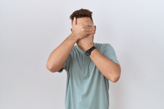 Hispanic man with beard standing over white background covering eyes and mouth with hands, surprised and shocked. hiding emotion